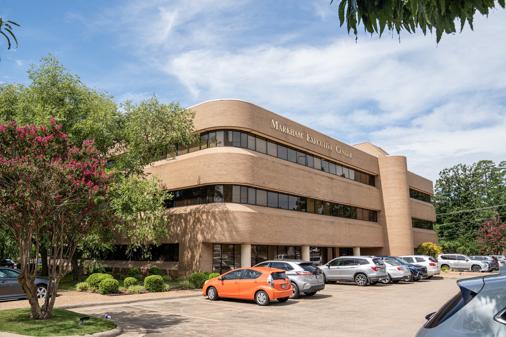 A low-angle, wide shot of the Markham Executive Center, a two-story, tan brick building with curved edges and rows of dark windows. An orange car is parked prominently in the paved lot in front of the building, with a blooming pink crape myrtle tree on the left.
