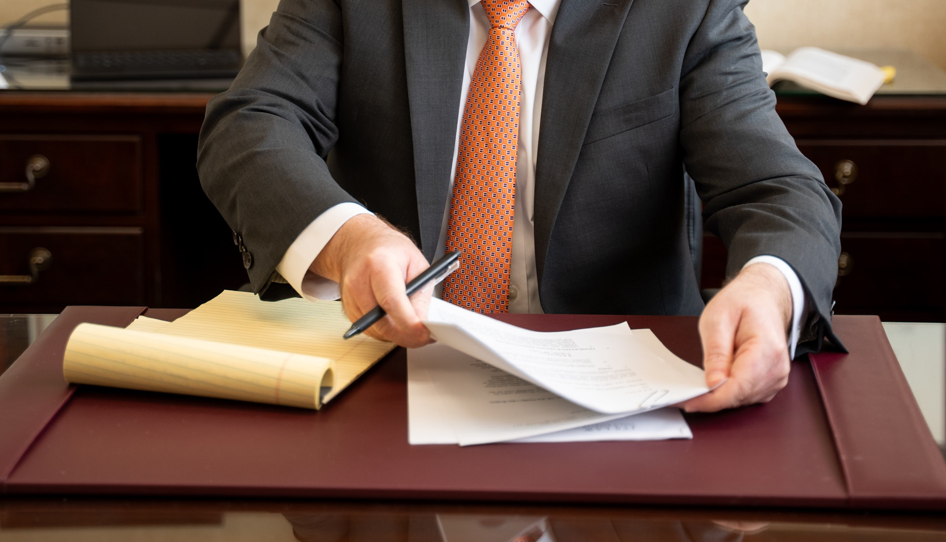 A cropped shot of a man wearing a dark suit and an orange patterned tie, sitting at a desk with a maroon leather blotter. He is holding a pen in one hand while using the other to flip through several documents on his desk, next to a legal pad.