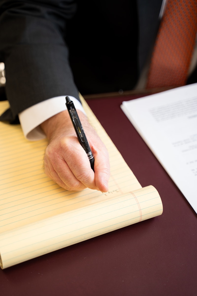 A close-up of a man's hand, wearing a dark suit and a white cuff, writing on a yellow legal pad with a black pen. The legal pad is on a desk with a maroon leather surface, with the man's orange tie visible in the background.