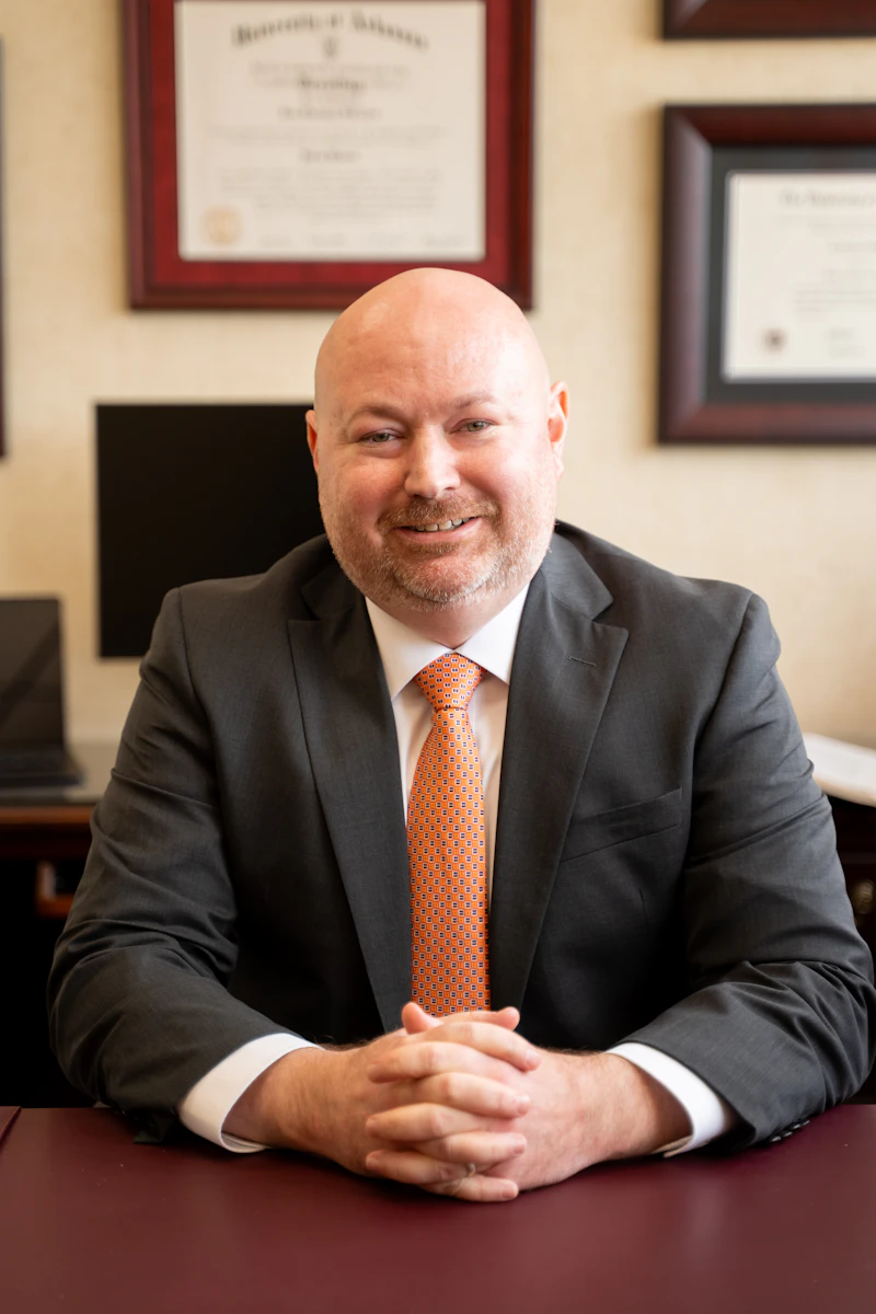 A professional headshot of a smiling Jon Shirron in a dark suit and an orange patterned tie. He is sitting at a desk with his hands clasped, and several framed awards are visible on the wall behind him.