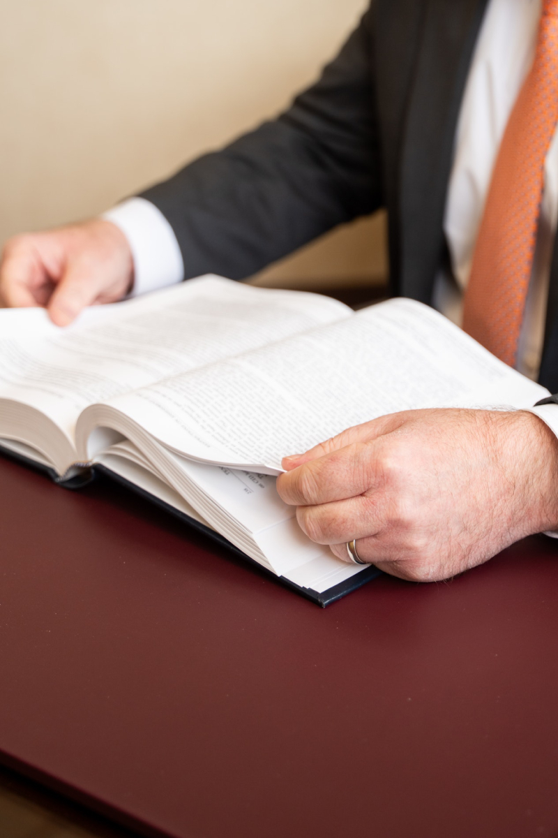 A close-up, cropped shot shows a man in a dark suit and orange tie sitting at a desk with a maroon leather surface. He is using both hands to hold and read a large, open book, with his fingers positioned to turn the pages.