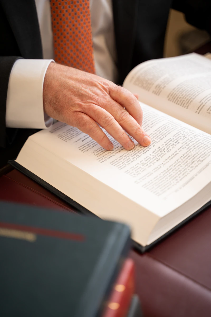 A close-up of a man's hands, wearing a dark suit and orange tie, as he turns a page of a large, open law book. A second, partially visible book is stacked on the maroon desk in the foreground.