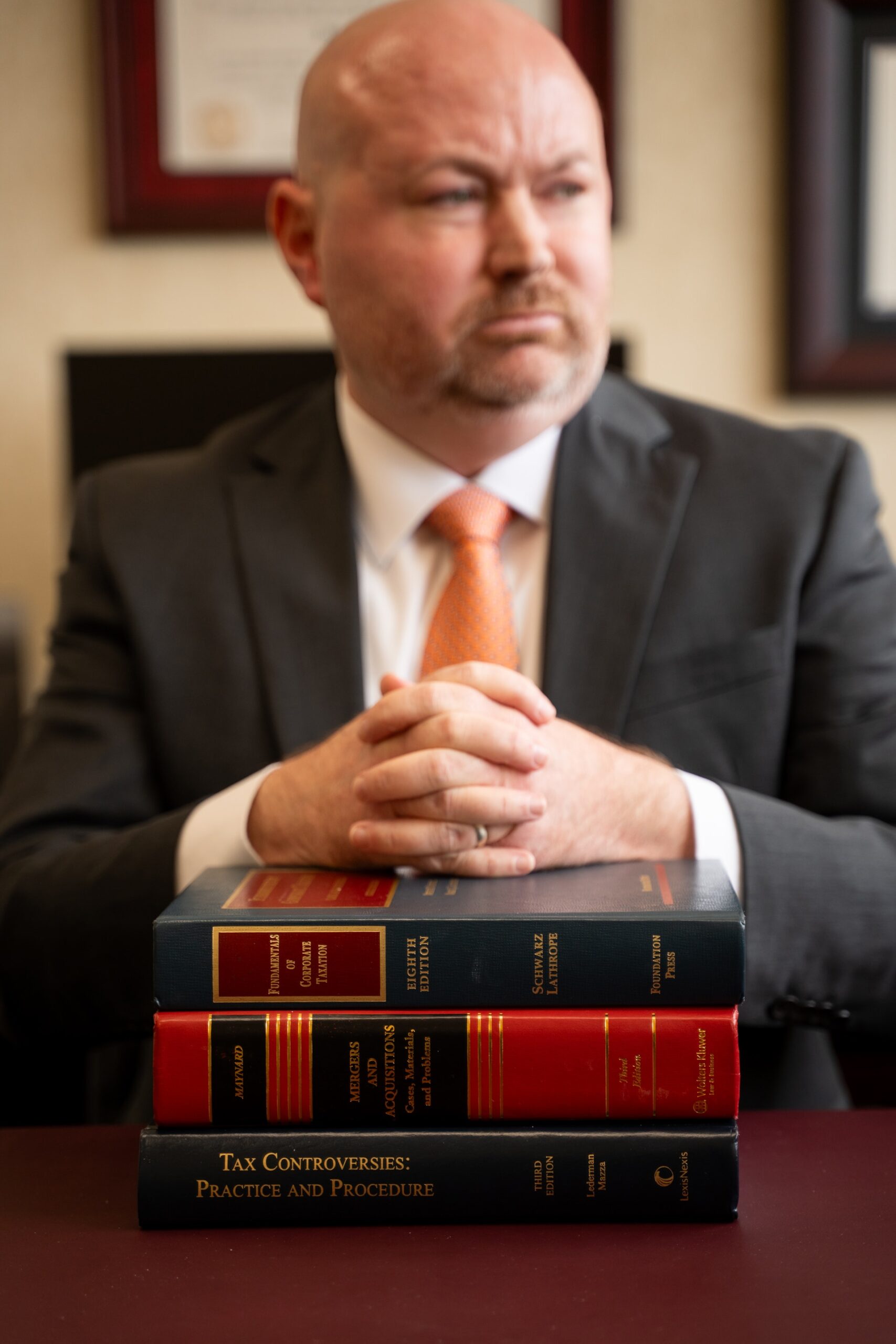 A man in a dark suit and an orange patterned tie sits behind a stack of three law books on a maroon desk. He has a serious expression and is resting his clasped hands on top of the books. Two framed documents are visible on the wall in the background.