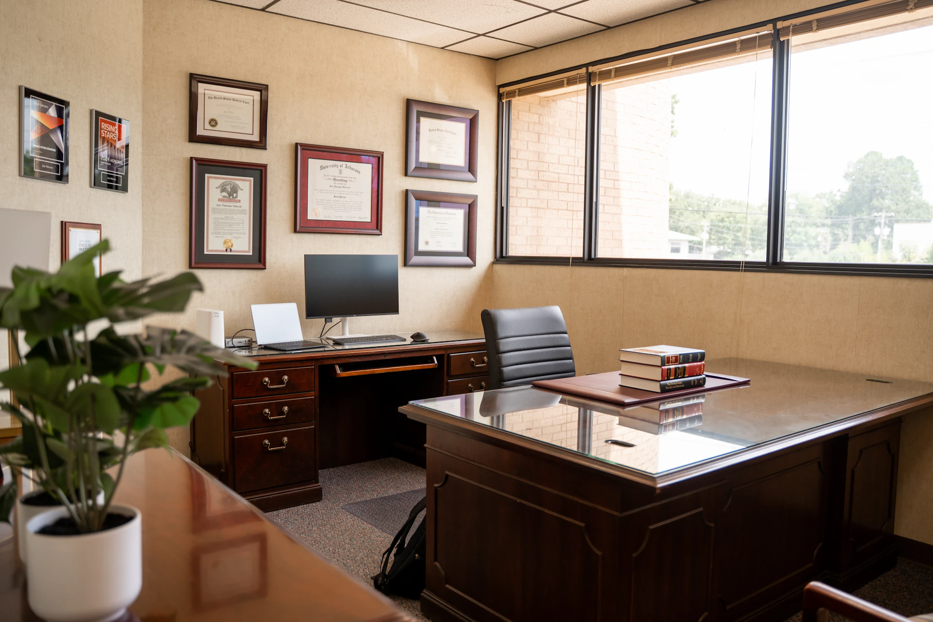 A clean and professional office features a large wooden desk with a stack of law books and a black leather chair. In the background, a smaller credenza holds a computer monitor and keyboard, while a large window lets in natural light and numerous awards and diplomas hang on the beige wall.