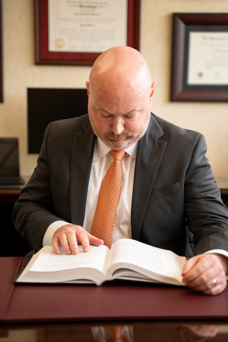 A professional headshot shows a Jon Shirron in a dark grey suit and an orange patterned tie. He is sitting at a desk with a maroon leather surface and is intently reading a large, open book. In the background, framed documents and certificates are visible on a beige wall.