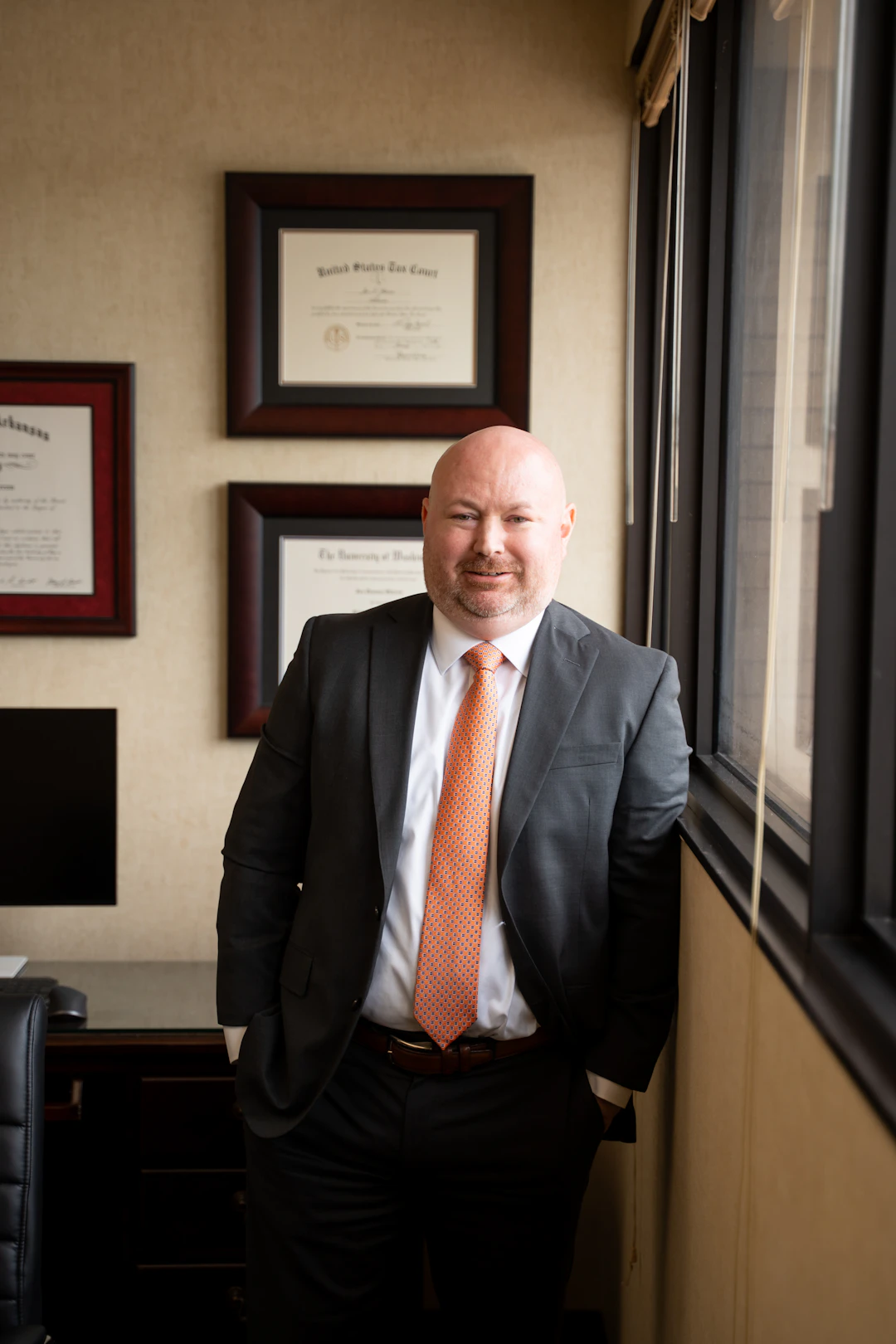 Smiling Jon Shirron in a dark suit and orange tie stands casually with his hands in his pockets, leaning against a wall next to a large window. In the background, three framed documents hang on a textured wall above a dark desk.