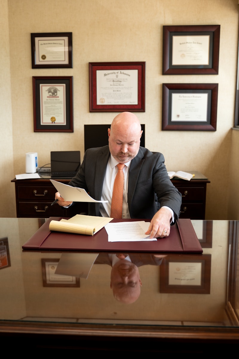 A clean and professional office features a large wooden desk with a stack of law books and a black leather chair. In the background, a smaller credenza holds a computer monitor and keyboard, while a large window lets in natural light and numerous awards and diplomas hang on the beige wall.
